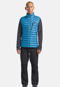 Gilet imbottito blu con motivi trapuntati orizzontali, collo alto e tasche con cerniera, indossato sopra una maglia a maniche lunghe grigio scuro e pantaloni neri.