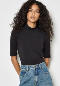 Top nero a maniche corte con collo alto, in tessuto liscio, design aderente, abbinato a pantaloni corti di jeans leggeri blu chiaro a vita alta con dettaglio dei bottoni.