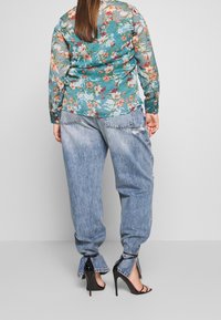 Blouse en teal transparent à imprimé floral avec des manches longues, associée à un jean loose-fit bleu clair avec des détails déchirés et des sandales à talons.
