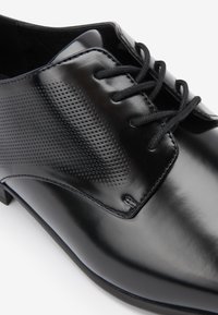 Black leather dress shoe with a glossy finish, featuring a perforated design on the side, round laces, and a low block heel.