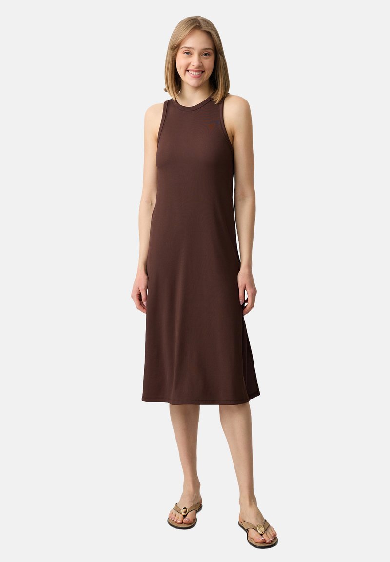 Jeune femme souriante, portant une robe midi sans manches marron et des sandales beiges sur un fond clair.