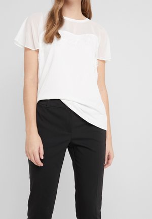 Frau trägt eine weiße, transparente Bluse mit kurzen Ärmeln und Spitzenverzierungen, kombiniert mit maßgeschneiderten schwarzen Hosen vor einem grauen Hintergrund.