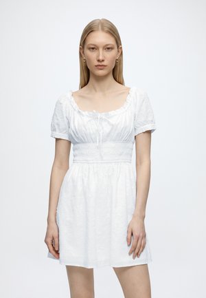 Junge Frau steht, trägt ein weißes Kleid mit kurzen Ärmeln, geraffter Taille und Rundhalsausschnitt vor einem einfarbigen hellen Hintergrund.
