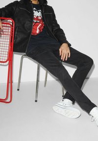 Person med svarta jeans, svart läderjacka och svart t-shirt med Rolling Stones sitter på en metallstol bredvid en röd metallstol.