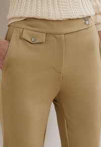 Μπεζ παντελόνι ψηλής μέσης με μπροστινή τσέπη και ασημένια κουμπιά, σε συνδυασμό με κρεμ πλεκτό πουλόβερ, χέρι στην τσέπη.