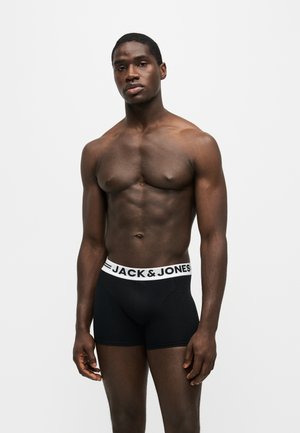 Homme musclé portant un boxer noir Jack & Jones, debout devant un fond blanc uni avec une expression neutre.