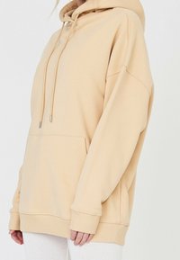 Béžová oversized mikina z měkké látky, s přední kapsou, nastavitelnými šňůrkami a žebrovanými manžetami. Hladká textura a volný střih.