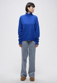 Plavi pleteni džemper s visokim ovratnikom, labavog kroja i dugih rukava, uparen sa svijetloplavim trapericama i smeđom obućom.