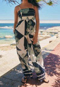 Femme portant une combinaison sans manches à jambes larges avec des motifs de feuilles tropicales noirs et beiges près d'une côte rocheuse par une journée ensoleillée.