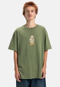 Oliwkowy bawełniany t-shirt o luźnym kroju, z nadrukiem graficznym przedstawiającym stos czaszek w białym i pomarańczowym kolorze na klatce piersiowej. Krótkie rękawy, okrągły dekolt.
