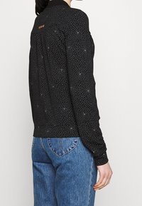 Svart långärmad topp med vita prickar, med krage och en rygghalsdetalj. Kombinerad med blå denimjeans.