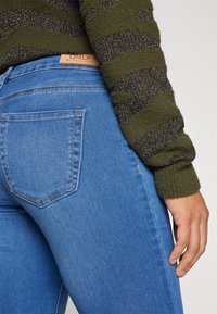 Jeans in denim blu con una vestibilità a vita media, caratterizzati da un classico design a cinque tasche e un lavaggio chiaro. Accompagnati da un maglione verde a trama.