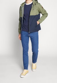 Grön och marinblå färgblockerad jacka med huva, bärs över en mörk marinblå skjorta, kombinerad med blå jeans och gula sneakers.