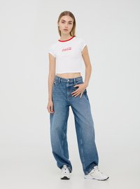 Maglietta bianca corta con colletto rosso e logo "Coca-Cola", abbinata a jeans in denim blu a taglio largo. Modello che indossa sneakers bianche.