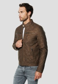 Hnedá motorkárska bunda z umelej kože so stojacím golierom, vreckami na zips a prešívaným detailom na ramenách, skombinovaná so svetlosivými džínami.