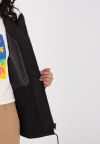 Veste zippée noire avec un intérieur lisse, cordon élastique à l'ourlet et une poche visible à l'intérieur, maintenue ouverte par une main.