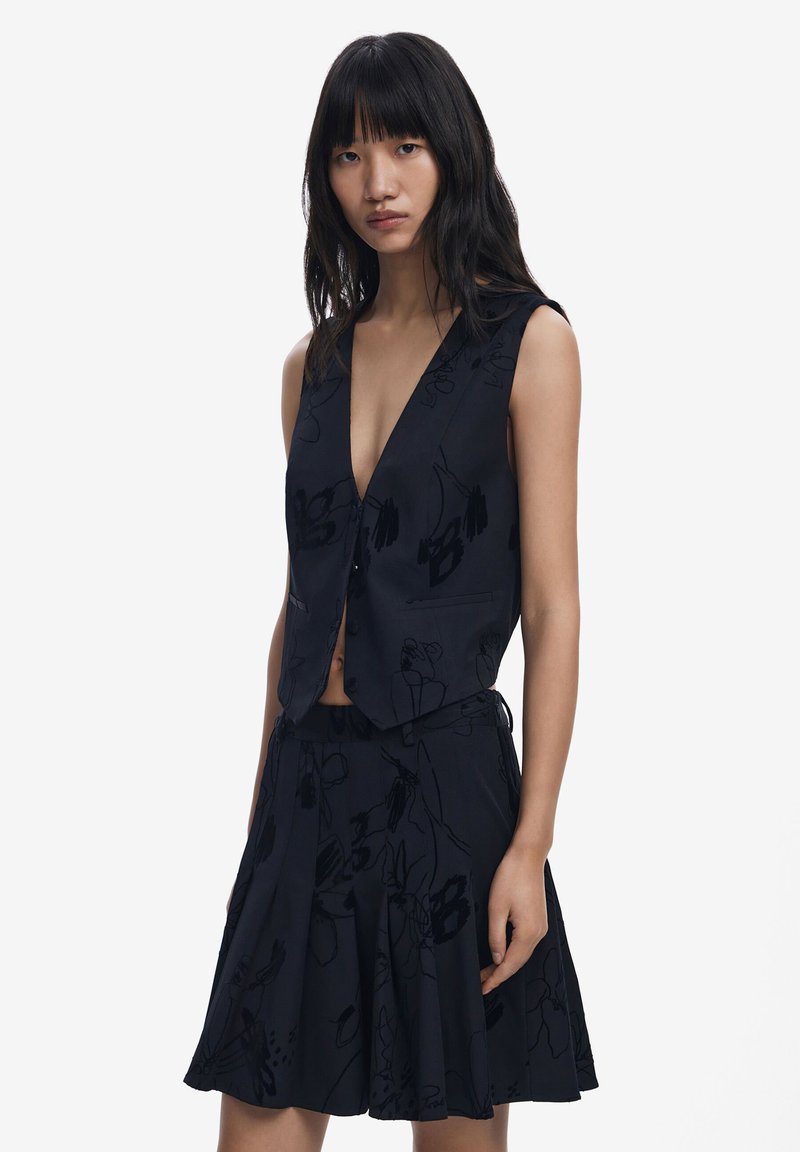 Svart blommönstrad väst med djup V-ringning och utan ärmar, tillsammans med en matchande utsvängd kjol. Tyget verkar slätt och lätt.