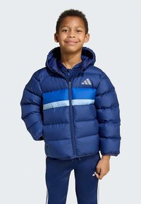 Berniukas stovi ir šypsosi, apsirengęs mėlynu pūkuotu džemperiu su kapišonu ir derančiomis mėlynomis sportinėmis kelnėmis su baltomis juostelėmis.