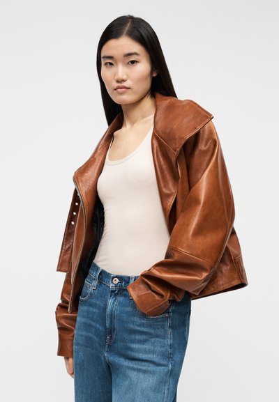 Brun skinnjacka med stor krage, dragkedja framtill och knappdetaljer, bärs över en beige linne och blå högmidjade jeans.