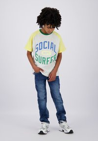 Maglietta grigia a maniche corte con maniche gialle, presenta la scritta blu e verde "SOCIAL SURFER", abbinata a jeans blu e sneaker bianche.