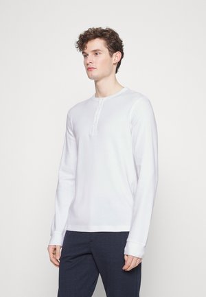 Langärmliges weißes Henley-Shirt aus weichem Baumwollstoff, mit Rundhalsausschnitt, Knopfleiste und gerippten Bündchen, kombiniert mit einer navyblauen Hose.