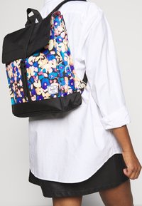 Sac à dos à motif floral avec un dessus et des bretelles en tissu noir. Forme rectangulaire avec une poche avant zippée. Logo de la marque à l'avant.