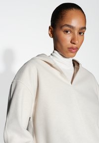 Hoodie oversize color crema in tessuto morbido con scollatura a V e cappuccio con cordini. Texture liscia, cuciture minime, abbinato a un dolcevita bianco.