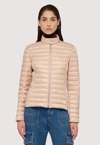 Světle růžová vypolstrovaná bunda s vysokým límcem, zipovým zapínáním a prošívanou strukturou. Nosená přes tmavě modré denimové kombinézy.