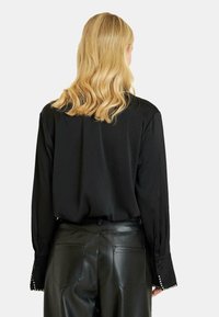 Blusa negra de manga larga con corte recto, textura suave y tachuelas decorativas en los puños. Combinada con pantalones de cuero negros ajustados.