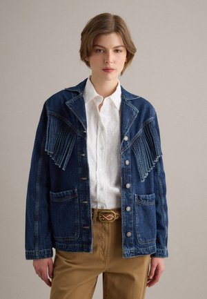 Jong persoon draagt een blauwe spijkerjas met franjes, een wit geborduurd overhemd en beige broek met een patroonriem, tegen een effen achtergrond.