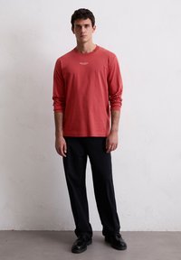 Lange mouwen, koraalkleurig T-shirt met een ronde hals en een subtiele merkl logo op de borst, gecombineerd met zwarte broeken en donkere schoenen.