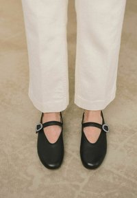 Zapatos de cuero negros con punta redonda, tira estilo Mary Jane y una hebilla decorativa de tono plateado, combinados con unos jeans de color crema.