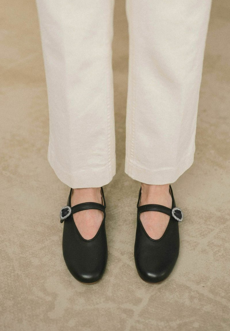 Zapatos de cuero negros con punta redonda, tira estilo Mary Jane y una hebilla decorativa de tono plateado, combinados con unos jeans de color crema.