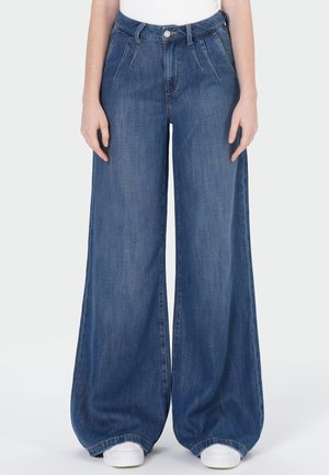 Femme portant un jean en denim bleu taille haute, jambes larges avec plis, associé à des baskets blanches, sur un fond clair uni.