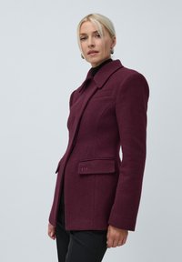 Woman with blonde hair wearing silver hoop earrings and a fitted deep purple wool blazer over black pants and top, standing against gray background.