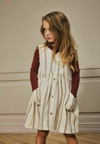 Flicka med långt hår iförd en krämfärgad klänning med bruna ränder och knappar, står med händerna i fickorna och tittar åt sidan.