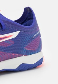 Bottines de course violettes avec un tissu à motifs, comportant un design en accents blancs et rouges. Semelle texturée avec des détails bleus et étiquetée "ULTRA".