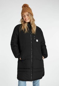 Sort lang pufferjakke med frontlynlås, sidelommer og et logoskilt; parret med en chunky, strikket brun hue.