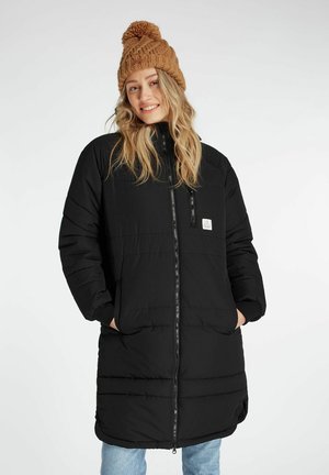 Zwarte lange pufferjas met een ritssluiting aan de voorkant, zijvakjes en een logopatch; gecombineerd met een chunky, gebreide bruine muts.