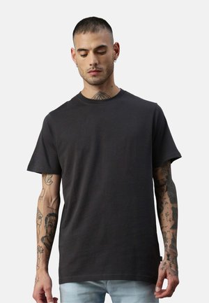 Junger Mann mit kurzen Haaren und Tattoos auf Armen und Hals, der ein schlichtes schwarzes T-Shirt und hellblaue Jeans trägt und nach unten blickt.