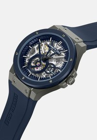 Granatowy zegarek automatyczny z przezroczystym szkieletowym cyferblatem, stalowymi akcentami, teksturowanym pierścieniem i gumowym paskiem. Posiada znakowanie marki Kenneth Cole.