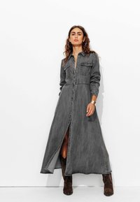 Robe longue en denim gris avec un design à boutons sur le devant, col, deux poches poitrine et une fente sur le côté, assortie à des bottines marron.
