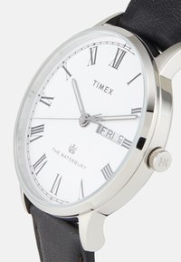Reloj redondo de tono plateado con esfera blanca, números romanos, correa de cuero negro, indicador de fecha y marca en la parte inferior.