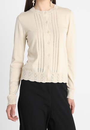Kobieta w kremowym swetrze na guziki z dzianiny z koronkowymi detalami na kołnierzu, mankietach, dolnej krawędzi oraz pionowymi rzędami z przodu, w połączeniu z czarnymi spodniami.