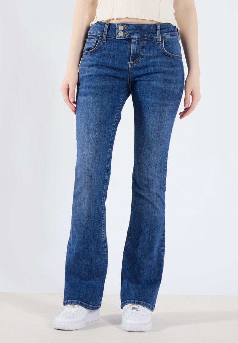 Jeans de campana azules hechos de denim, con cierre de botones, diseño de cinco bolsillos y un ligero desgaste. Llevados con zapatillas blancas.