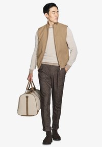 Tan vest over en lys sweater, brune strukturerede bukser, brune ankelstøvler, bærende en beige duffel taske med brune detaljer.