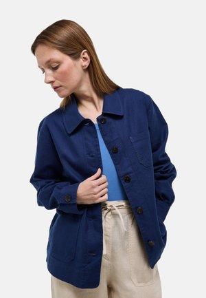 Femme en pantalon beige et chemise bleue portant une veste bleu foncé boutonnée, regardant vers le bas et ajustant la veste d'une main.