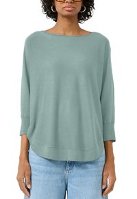 Kobieta w luźnym, jasnoniebiesko-zielonym swetrze z dekoltem w łódkę, z rękawami do trzech ćwiartek, oraz jasnoniebieskich dżinsach, skróconych do podbródka i w talii.