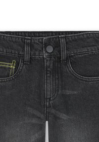 Gros plan sur un jean en denim noir montrant le bouton avant, les passants de ceinture et le petit détail de poche cousue en jaune sur le côté gauche.