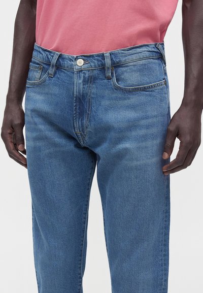Homme portant un jean en denim bleu moyen et une chemise rose rentrée, mains détendues sur les côtés, visible de la taille à mi-cuisse.
